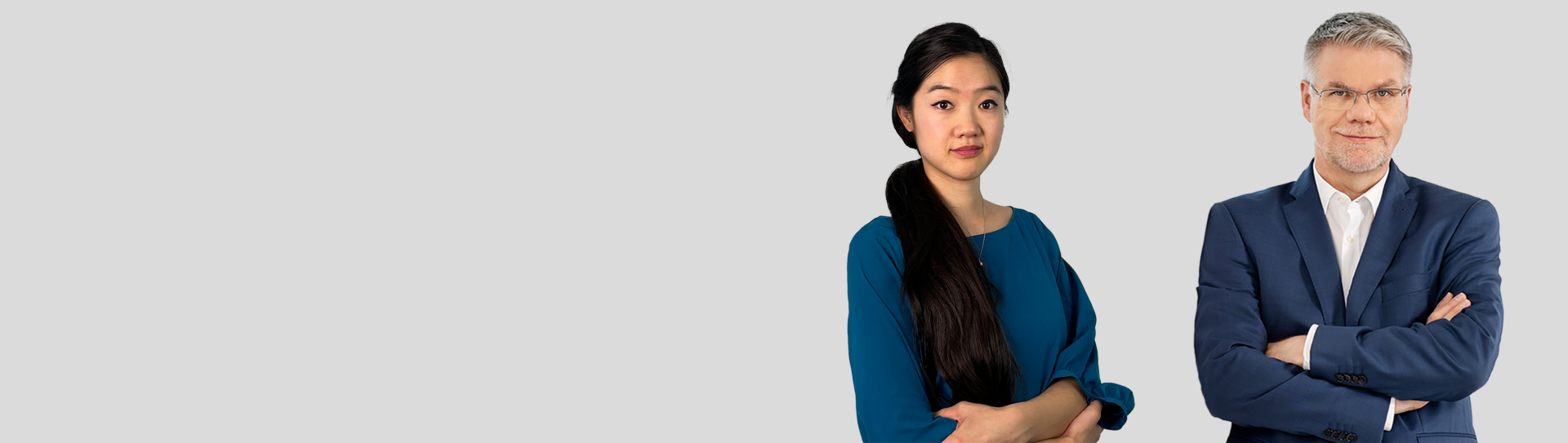 Das Bild zeigt zwei Personen in formeller Kleidung; eine Frau in einem blauen Kleid links und einen Mann in einem blauen Anzug rechts. Beide Personen haben die Arme gekreuzt.